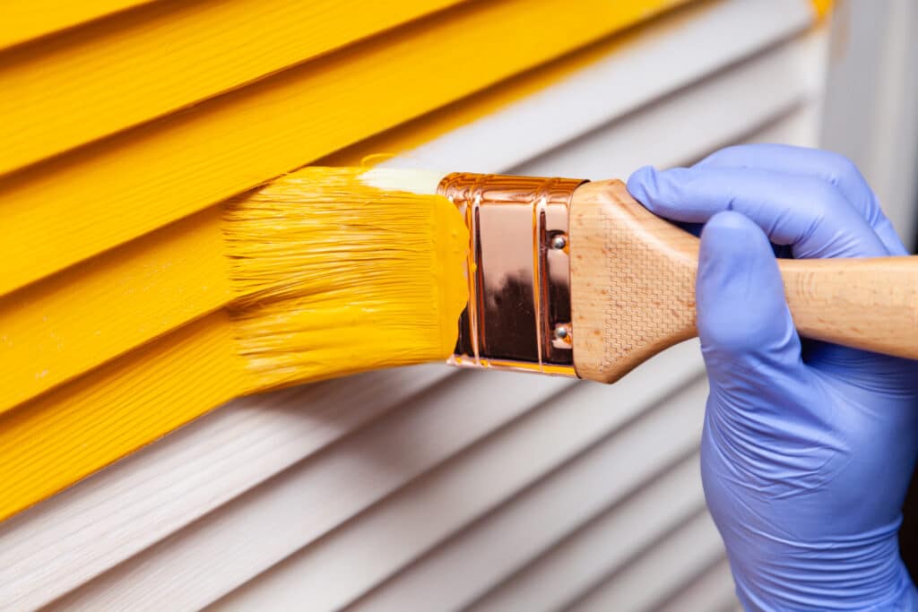 Main gantée de bleu peignant des persiennes en bois en jaune vif avec un pinceau.
