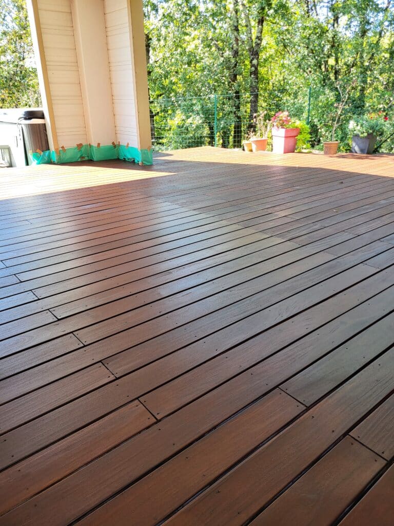 Terrasse en bois brun avec zones de soleil et d'ombre, vue sur un jardin verdoyant. Murs protégés et spa visibles à gauche.