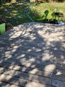 Terrasse en bois patinée sous des ombres d'arbres, bordant un jardin verdoyant avec un étendoir à linge et des plantes en pot.