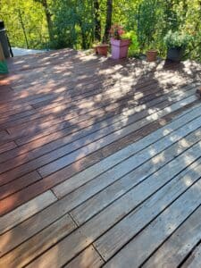 Terrasse en bois montrant le contraste entre des lattes vieillies et d'autres fraîchement traitées. Verdure, arbres et pots de fleurs.